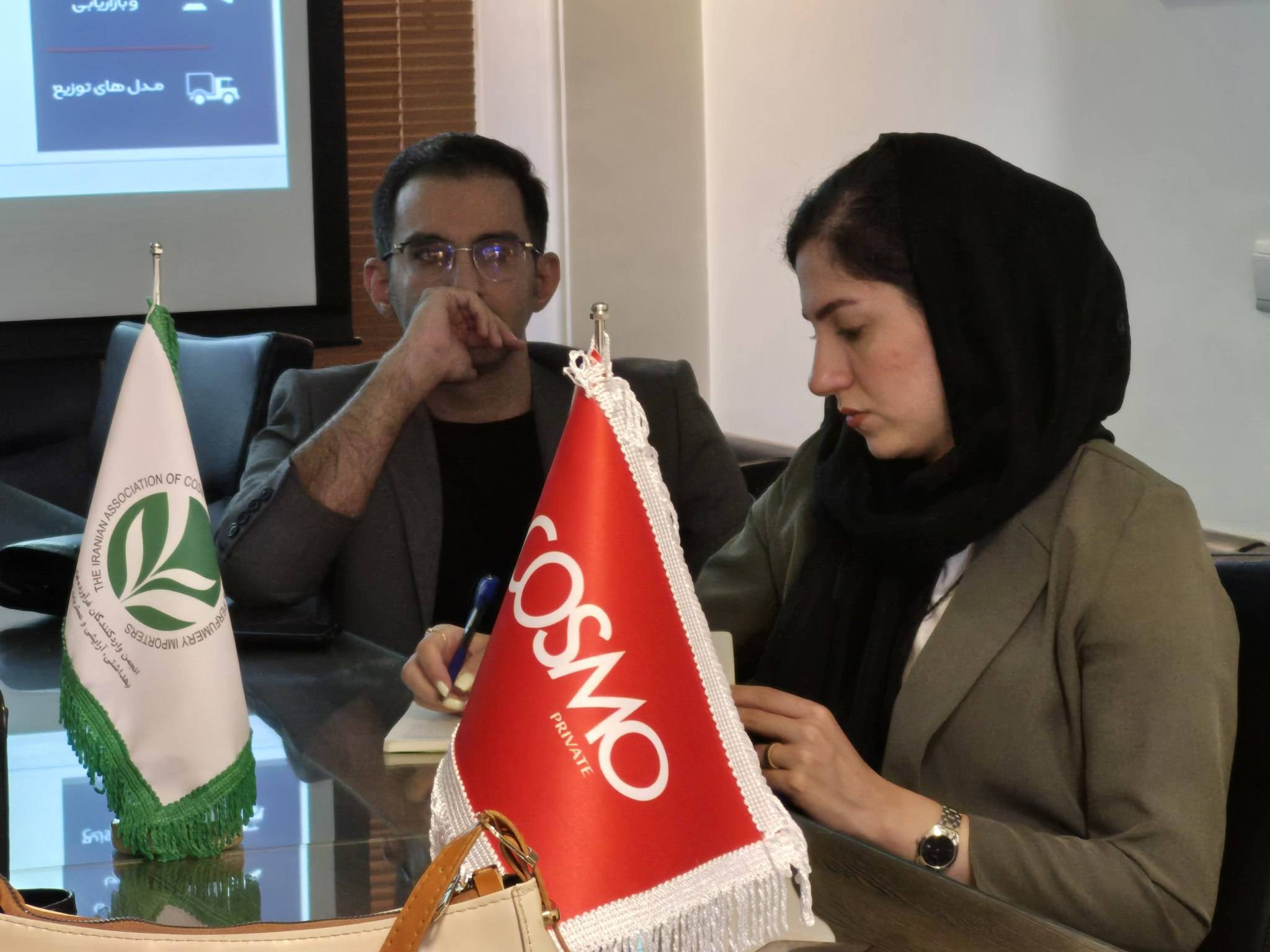 شرکت‌کنندگان در کارگاه تولید بدون کارخانه، در حال بحث و یادداشت‌برداری، با پرچم‌های COSMO و انجمن واردکنندگان محصولات کشاورزی در پس‌زمینه.